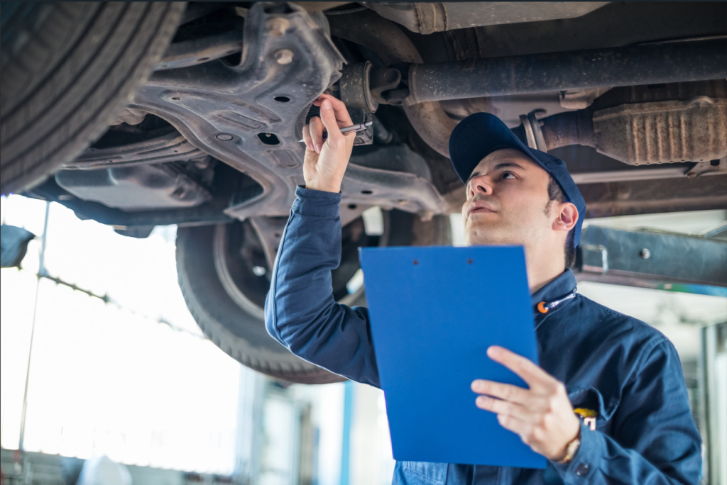 A man underneath a vehicle with a blue clipboard inspecting the under side of a vehicle near Lawrence, MA.